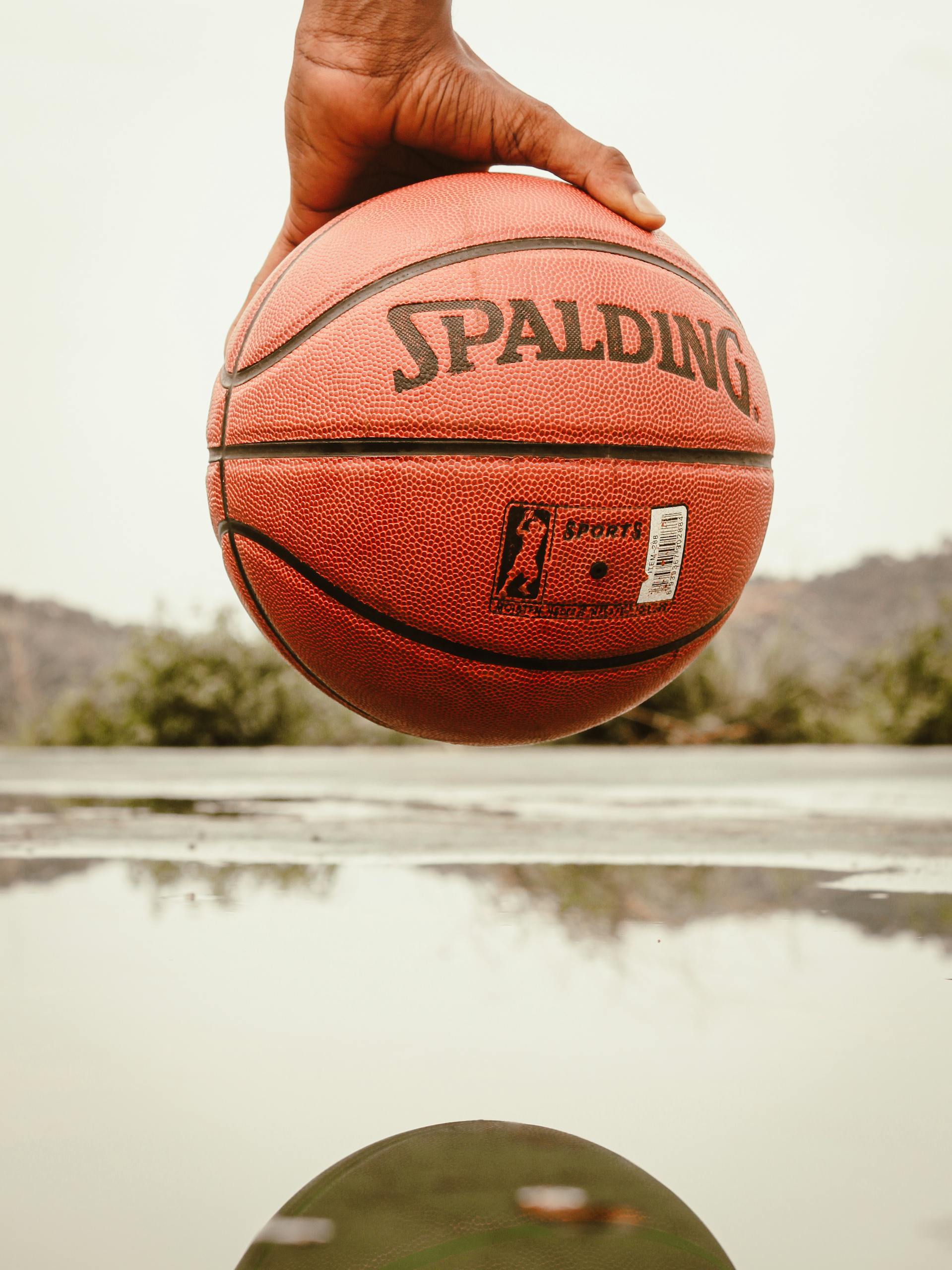 A close-up shot of a hand holding a basketball outdoors near a reflective puddle.