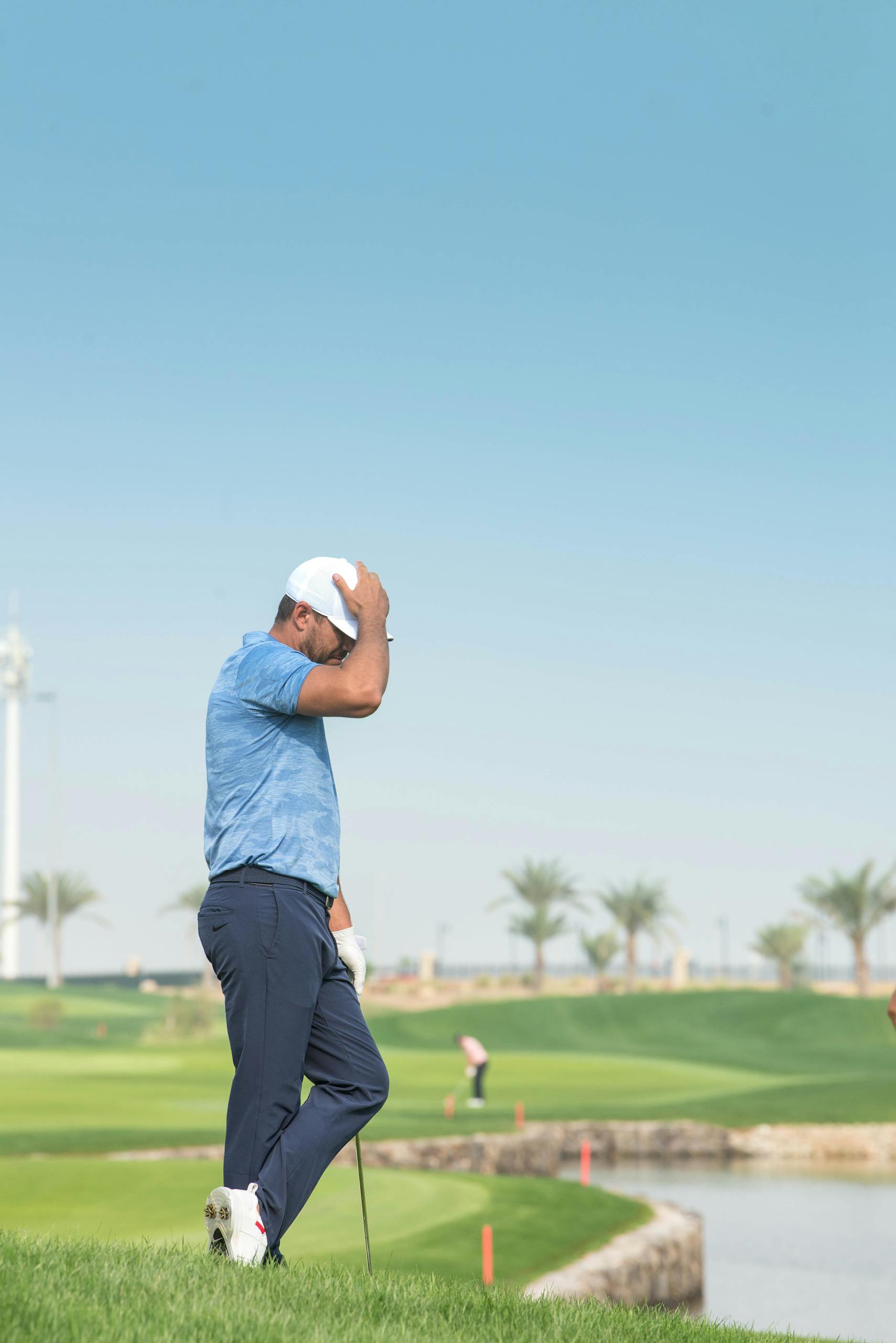 A golfer in a blue shirt stands disappointed on a sunny golf course, contemplating a missed shot.