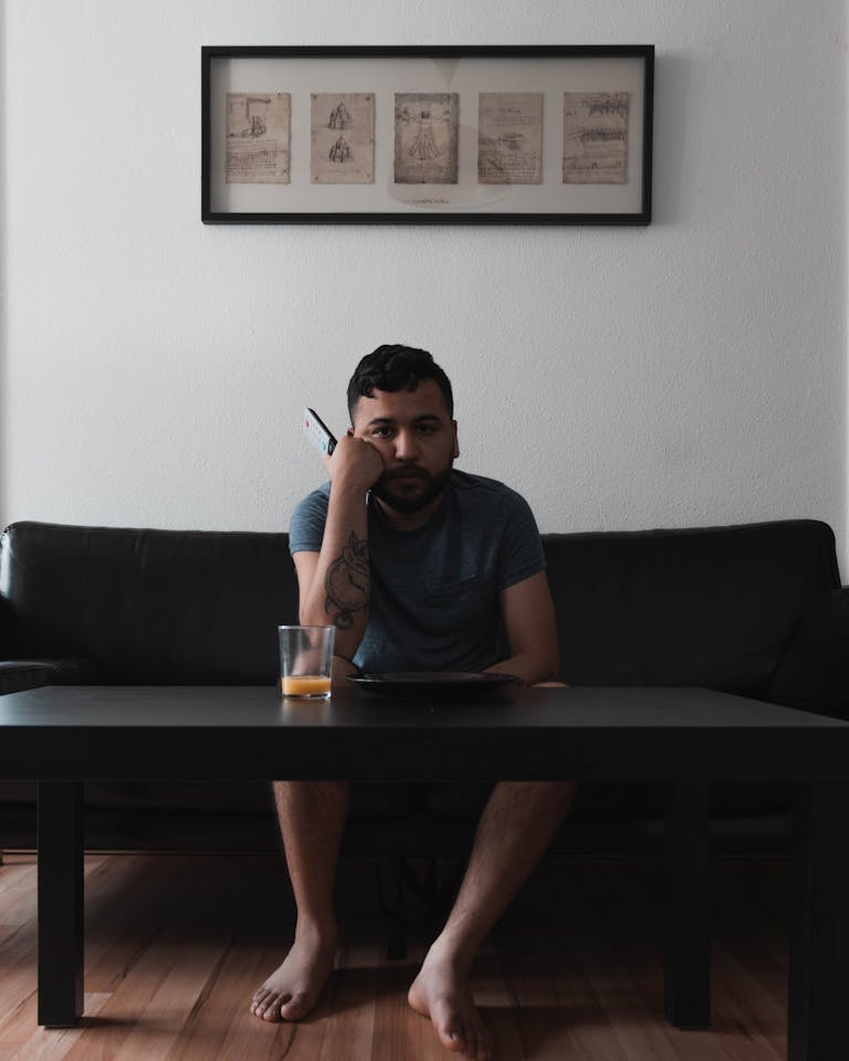 An adult man sitting on a sofa indoors holding a remote, juice on table.