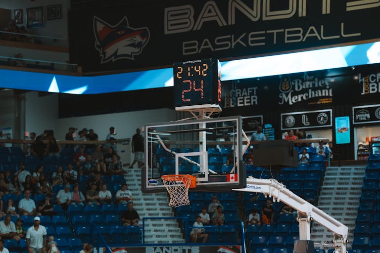 Close-up of a basketball hoop inside a stadium with a scoreboard and audience in the background.