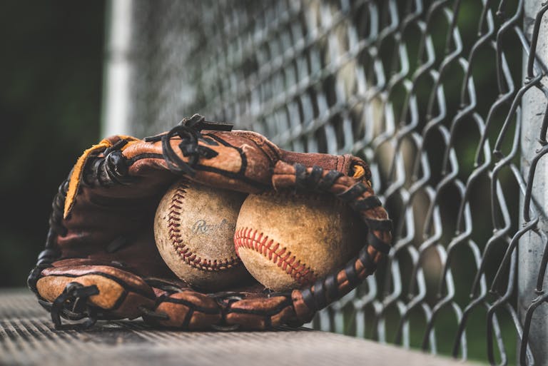 Close-up of a leather glove holding two vintage baseballs, resting near a chain-link fence.