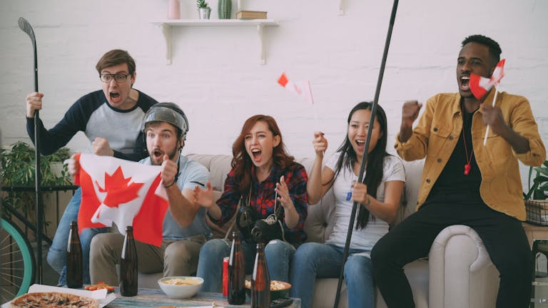 Diverse friends cheer for Canada with flags and hockey gear in a lively room.