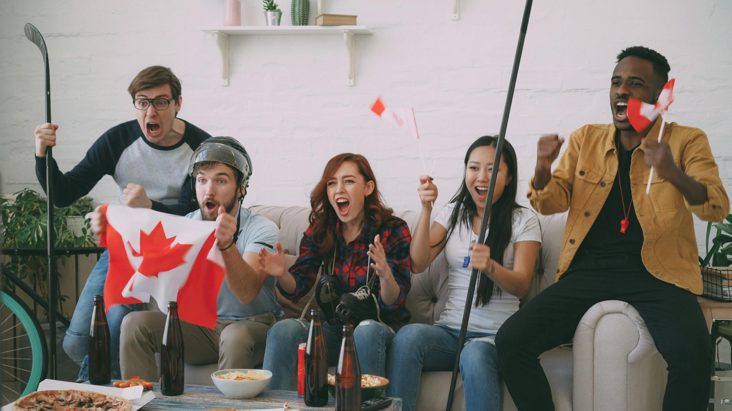 Diverse friends cheer for Canada with flags and hockey gear in a lively room.