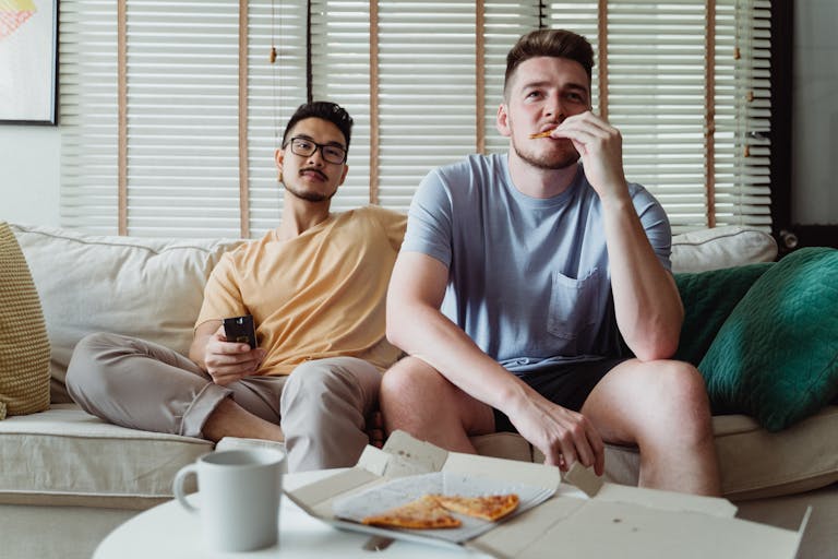 Two young men enjoying pizza and TV, sitting on a comfortable couch indoors.