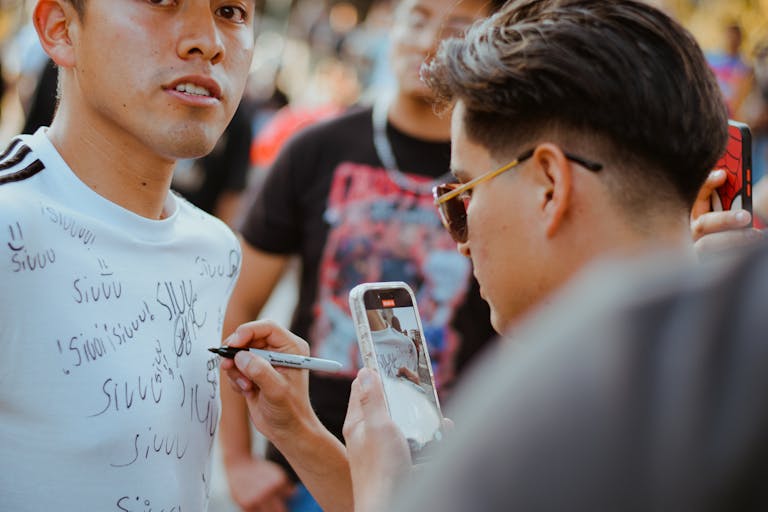 Young adults signing t-shirts at an outdoor event, capturing the moment with smartphone.
