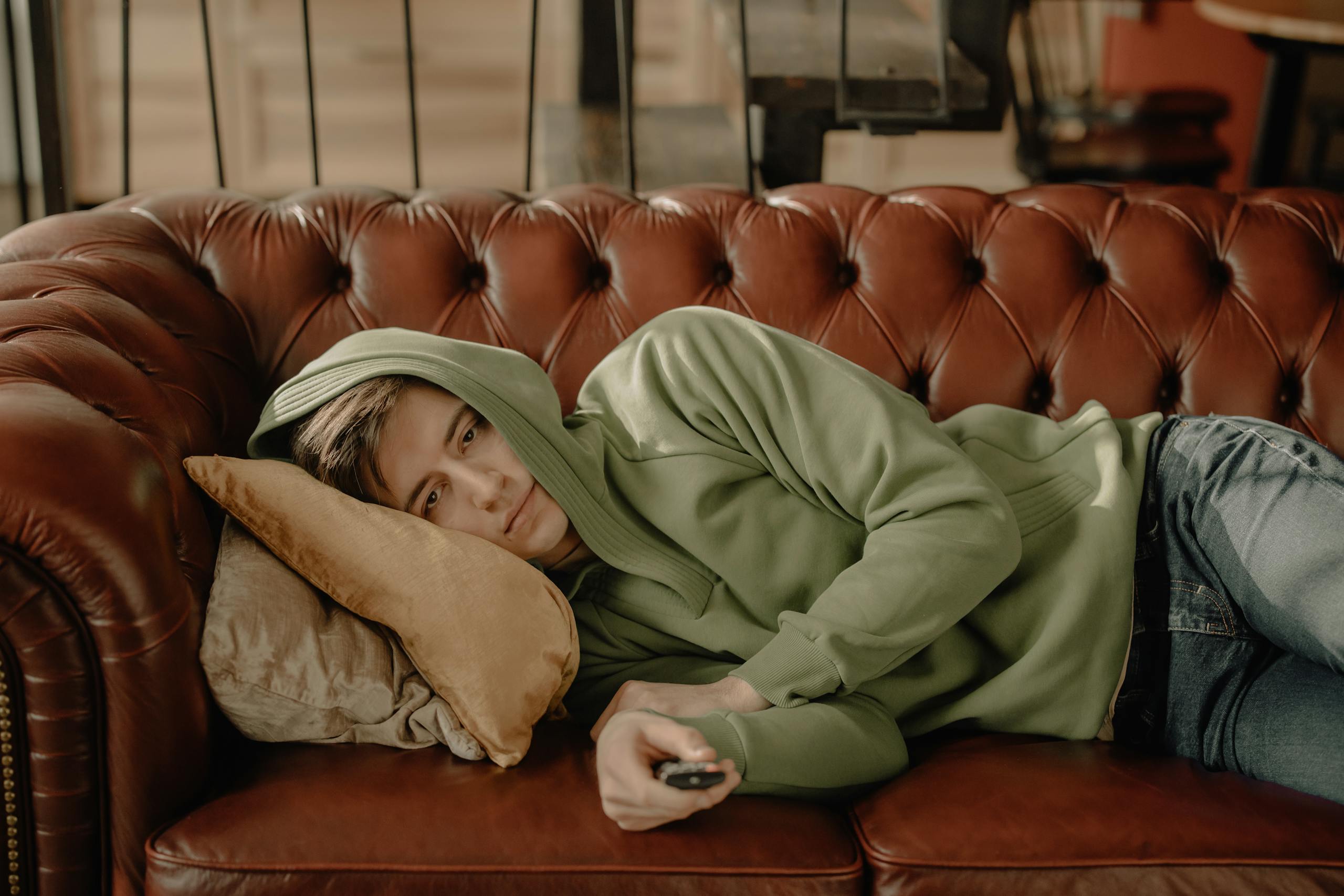 Young man in green hoodie relaxing on a leather sofa at home holding a remote.