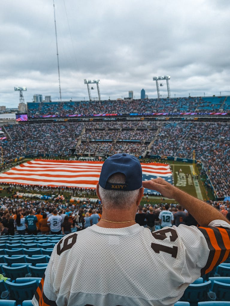 A fan in a sports jersey salutes at a lively football game with a giant American flag.