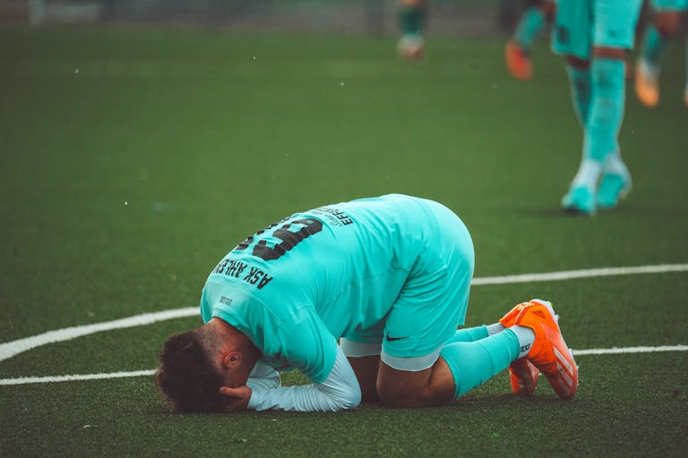 A soccer player kneels in despair on a green artificial turf sports field.