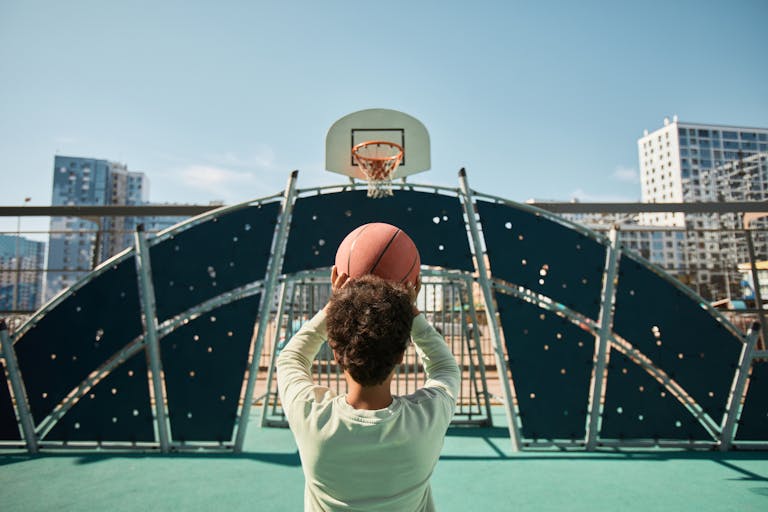 A young boy aims to shoot a basketball on an outdoor urban court, under a clear blue sky.