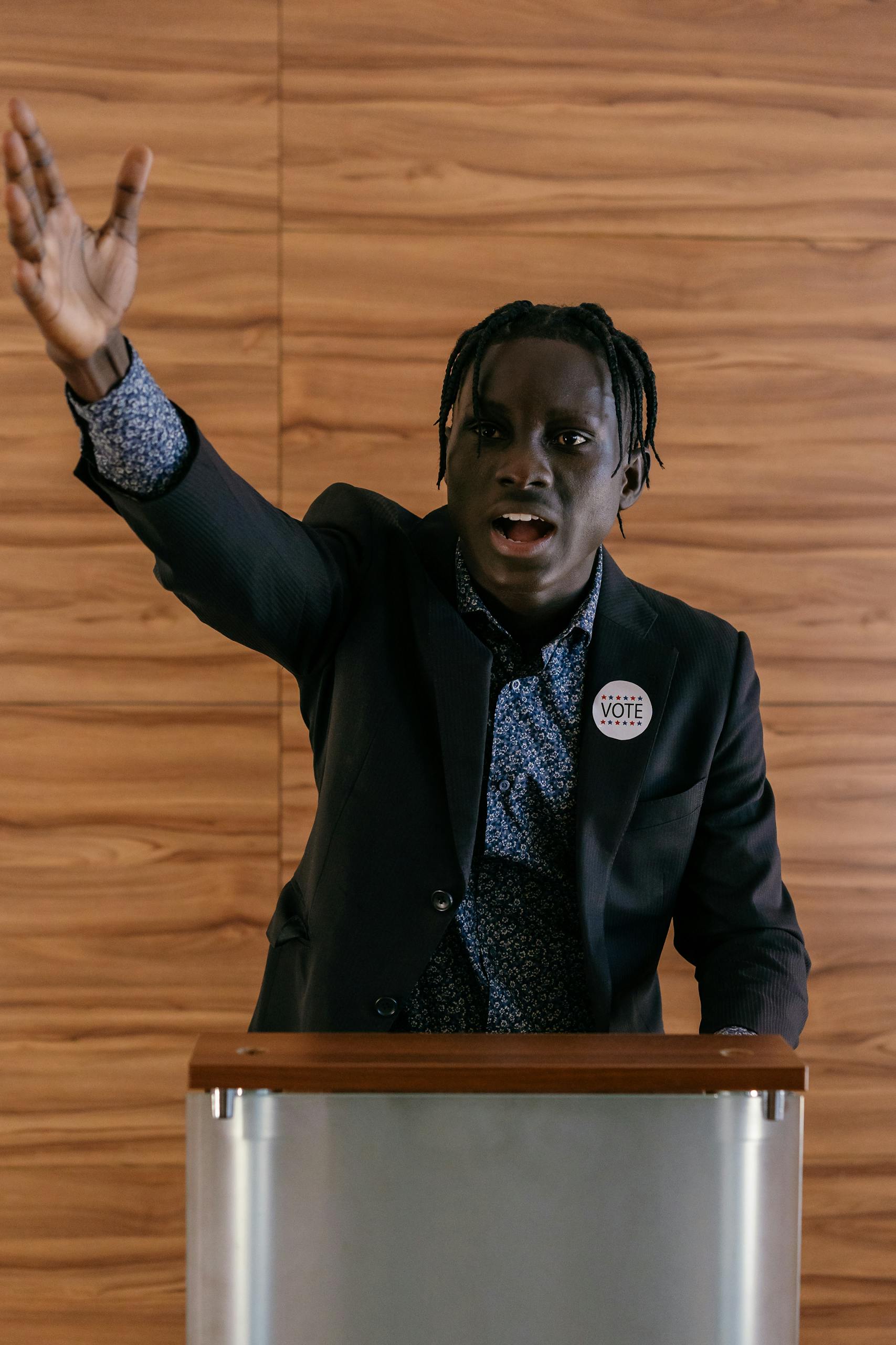 Black male politician delivering a speech with expressive hand gesture at a podium indoors.