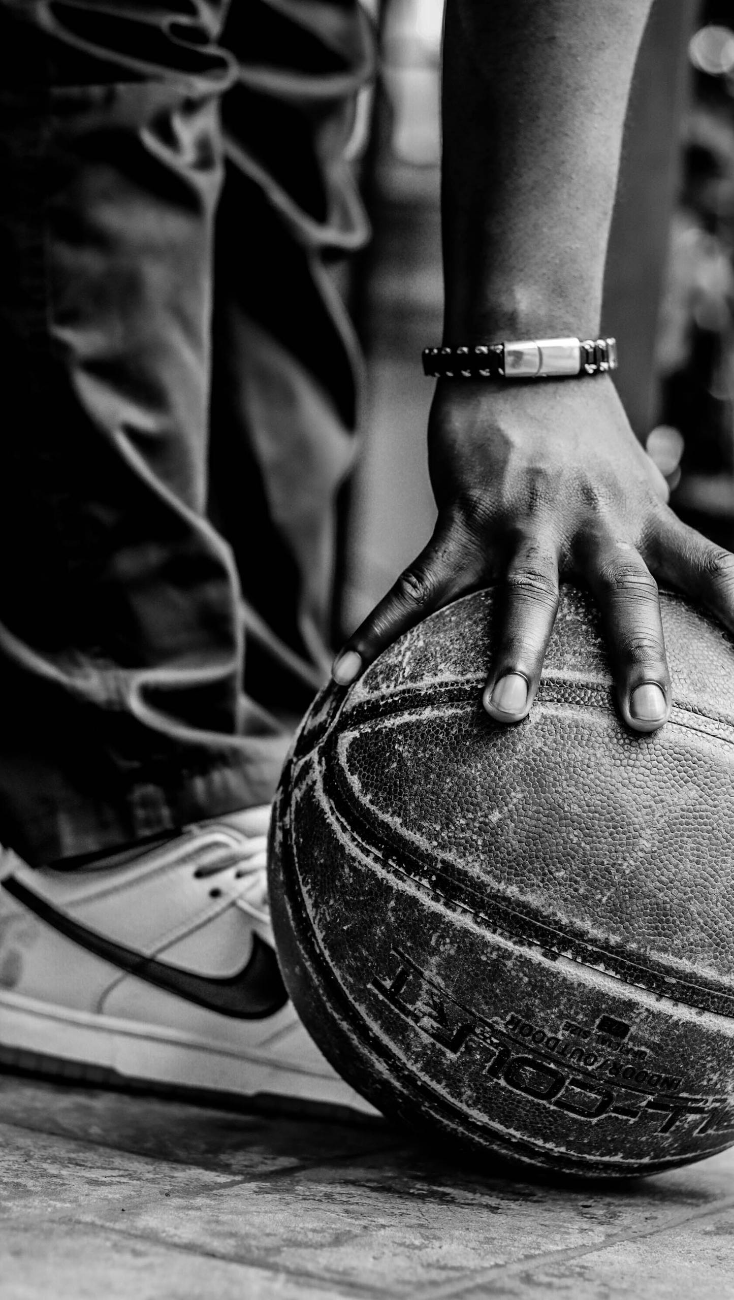 Monochrome image of a basketball player clutching a ball, showcasing details of fashion footwear and wrist bracelet.