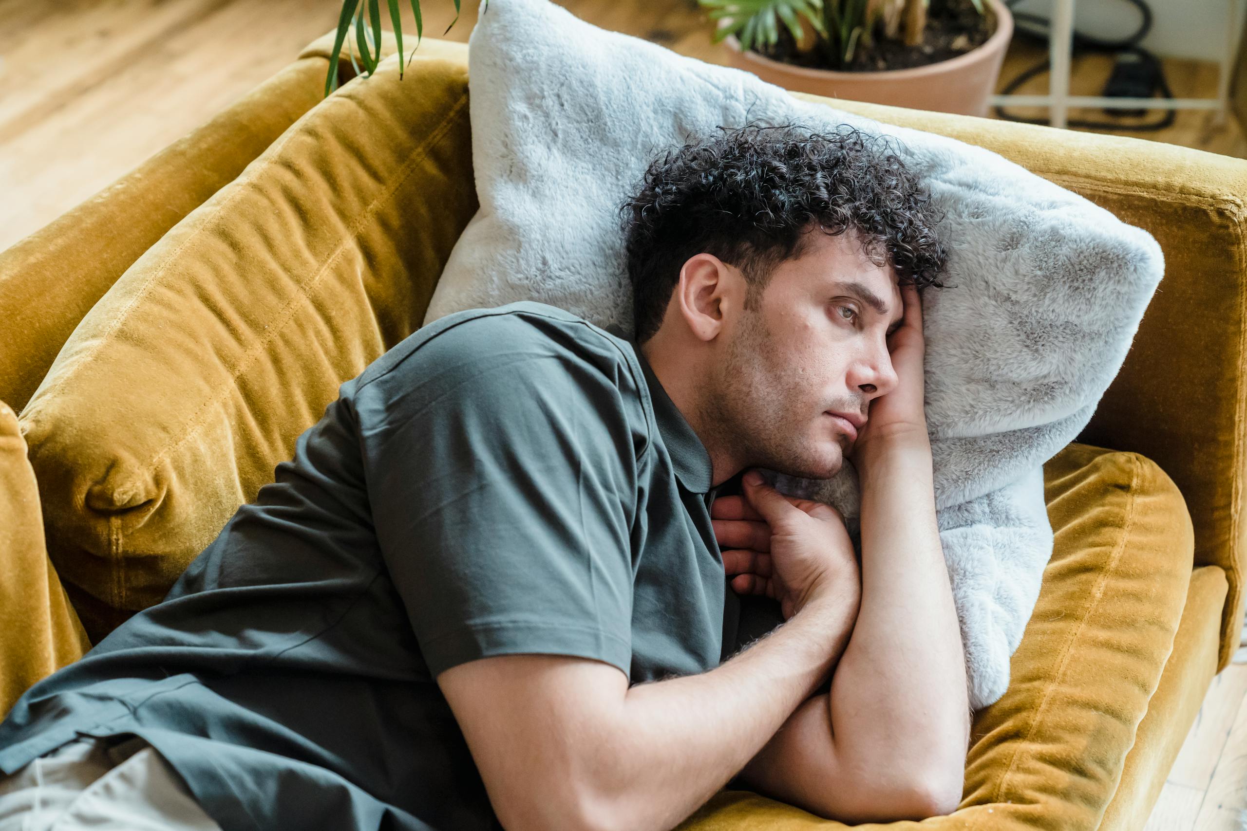 Thoughtful young man relaxing on a warm-toned couch with a soft pillow, conveying calm and introspection.