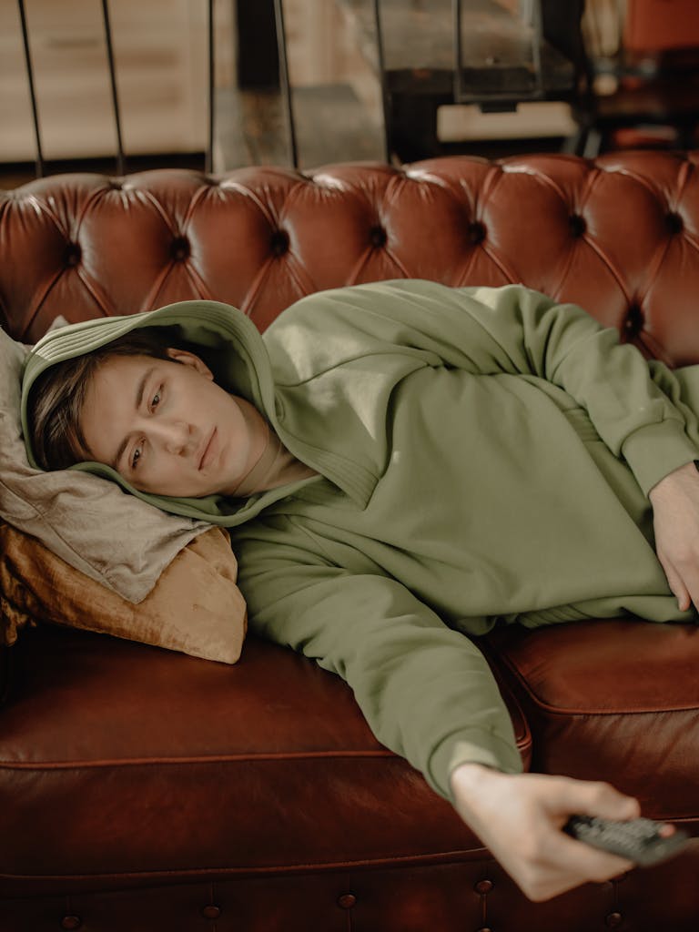Young man in green hoodie relaxing on a leather sofa with a remote control, indoors.