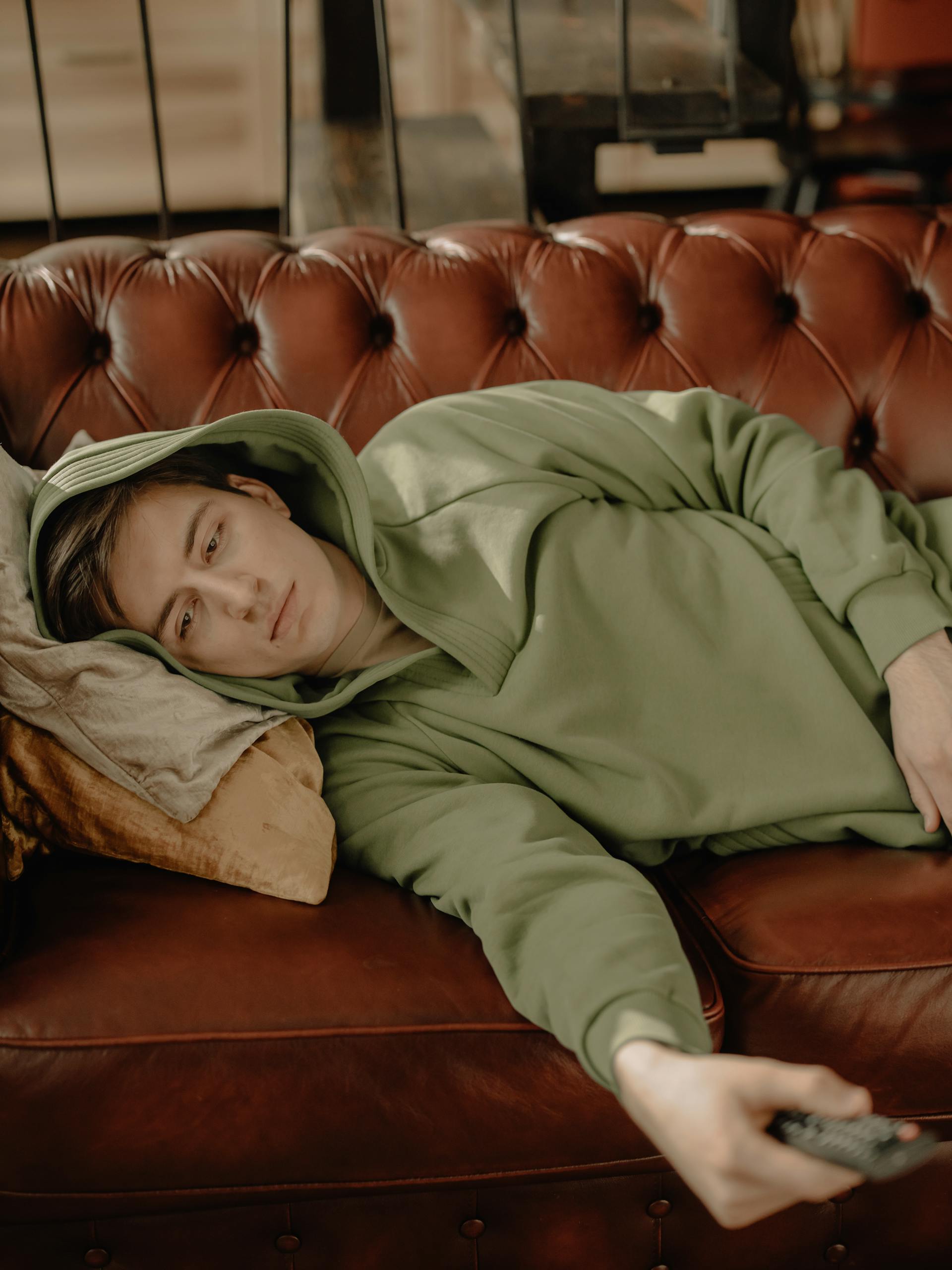 Young man in green hoodie relaxing on a leather sofa with a remote control, indoors.