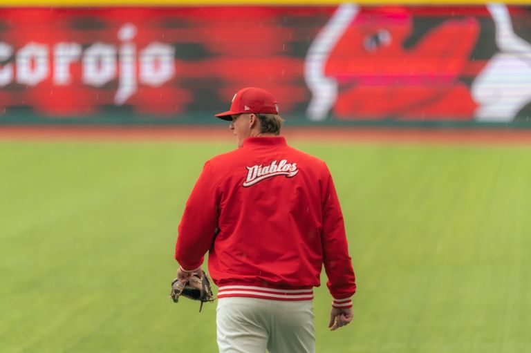 A baseball player wearing a red jacket and hat stands on a grassy field during a game.