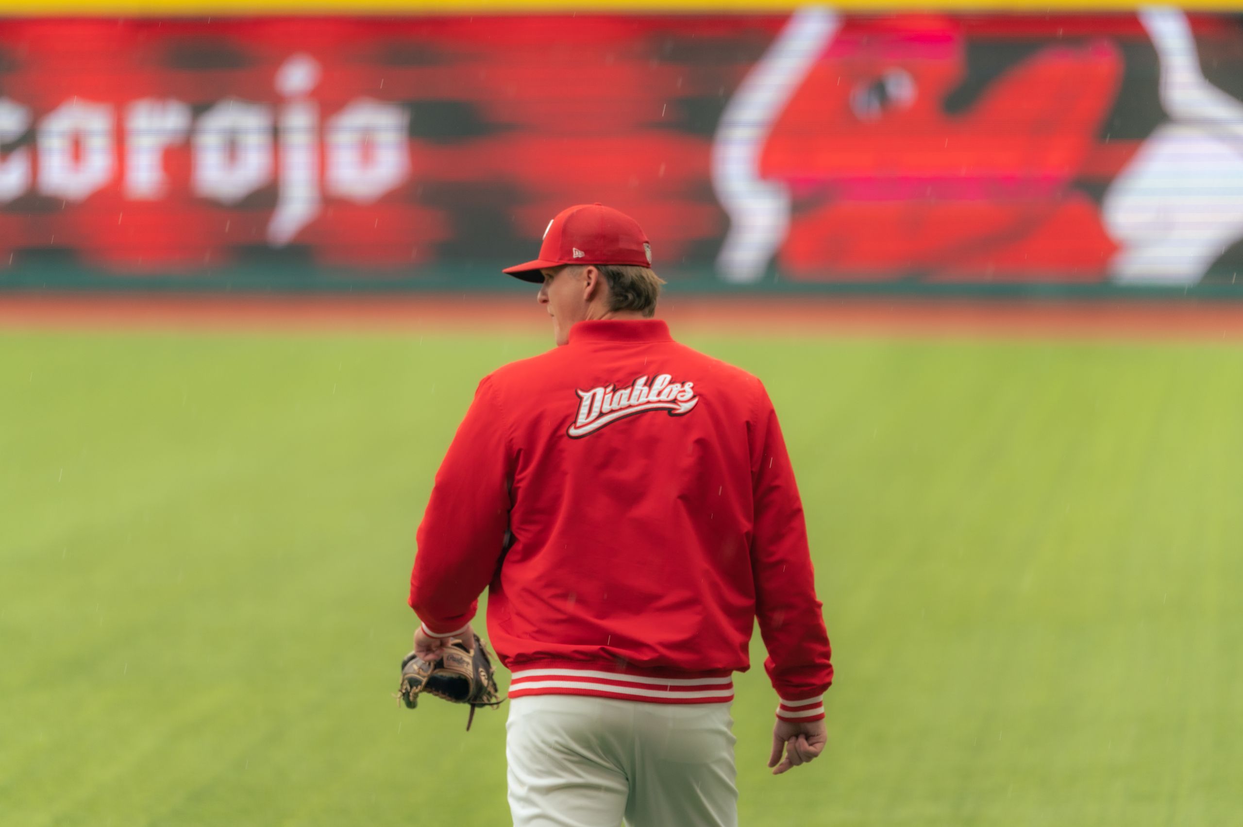 A baseball player wearing a red jacket and hat stands on a grassy field during a game.