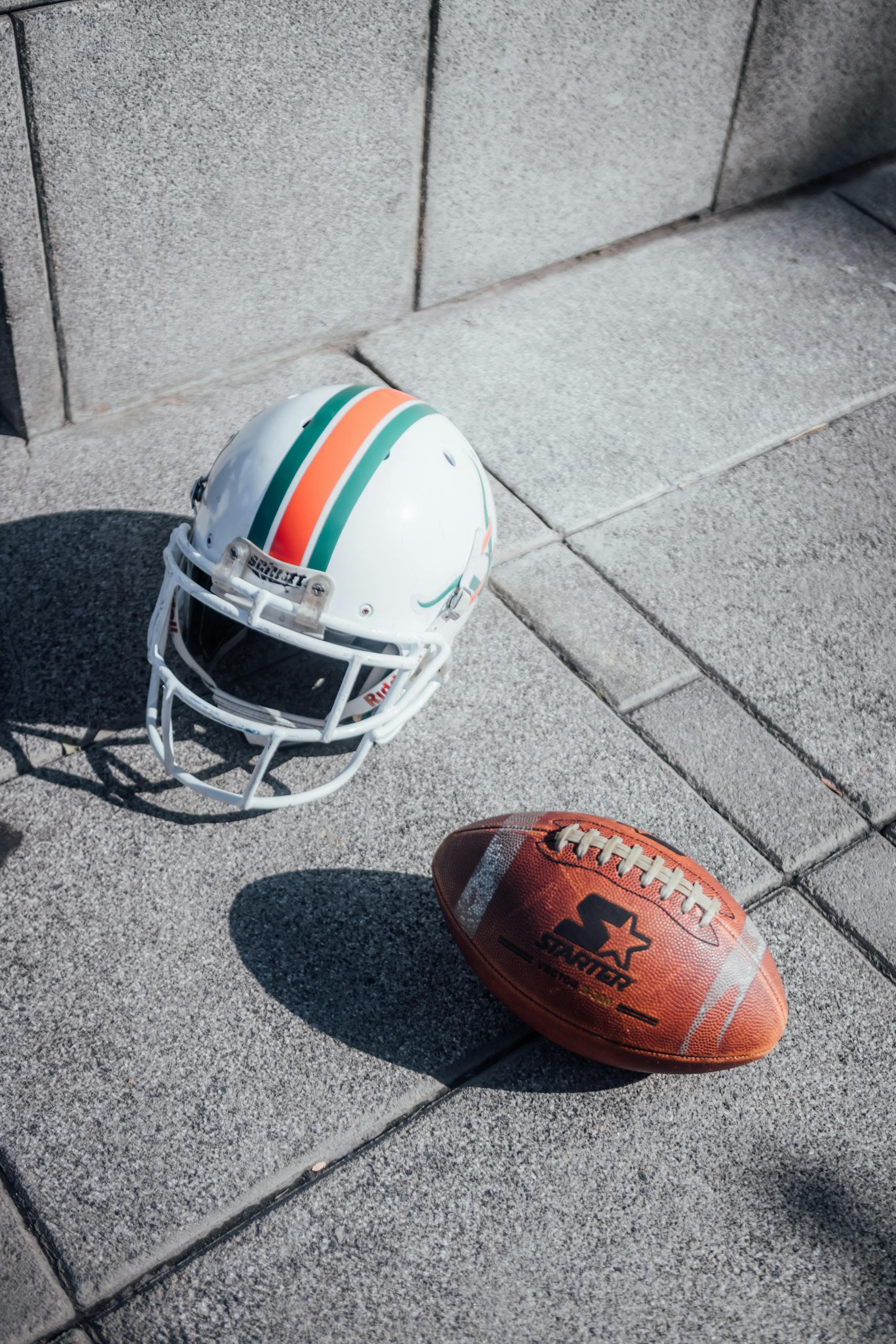 A close-up shot of an American football and helmet on a city sidewalk under sunlight.