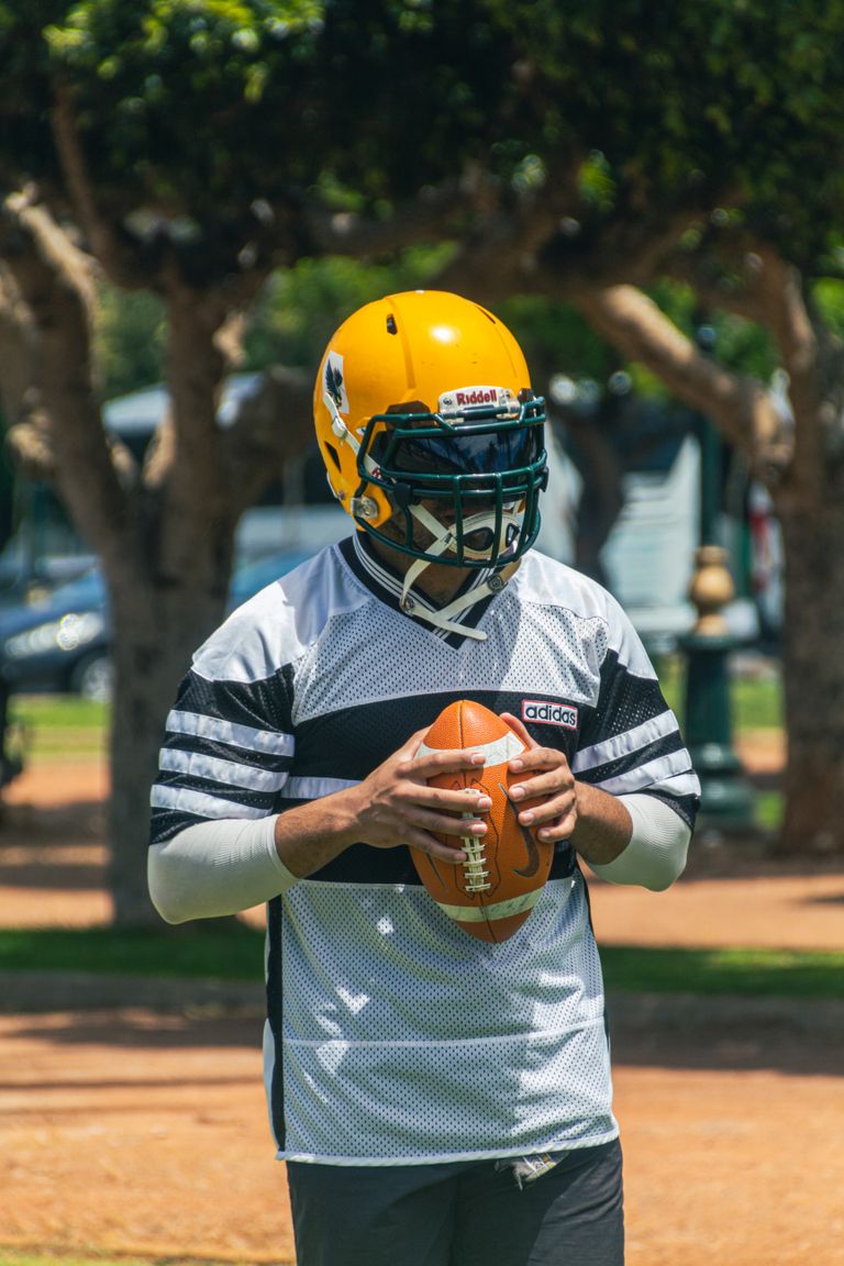 A football player in full gear holding a ball during practice outdoors on a sunny day.