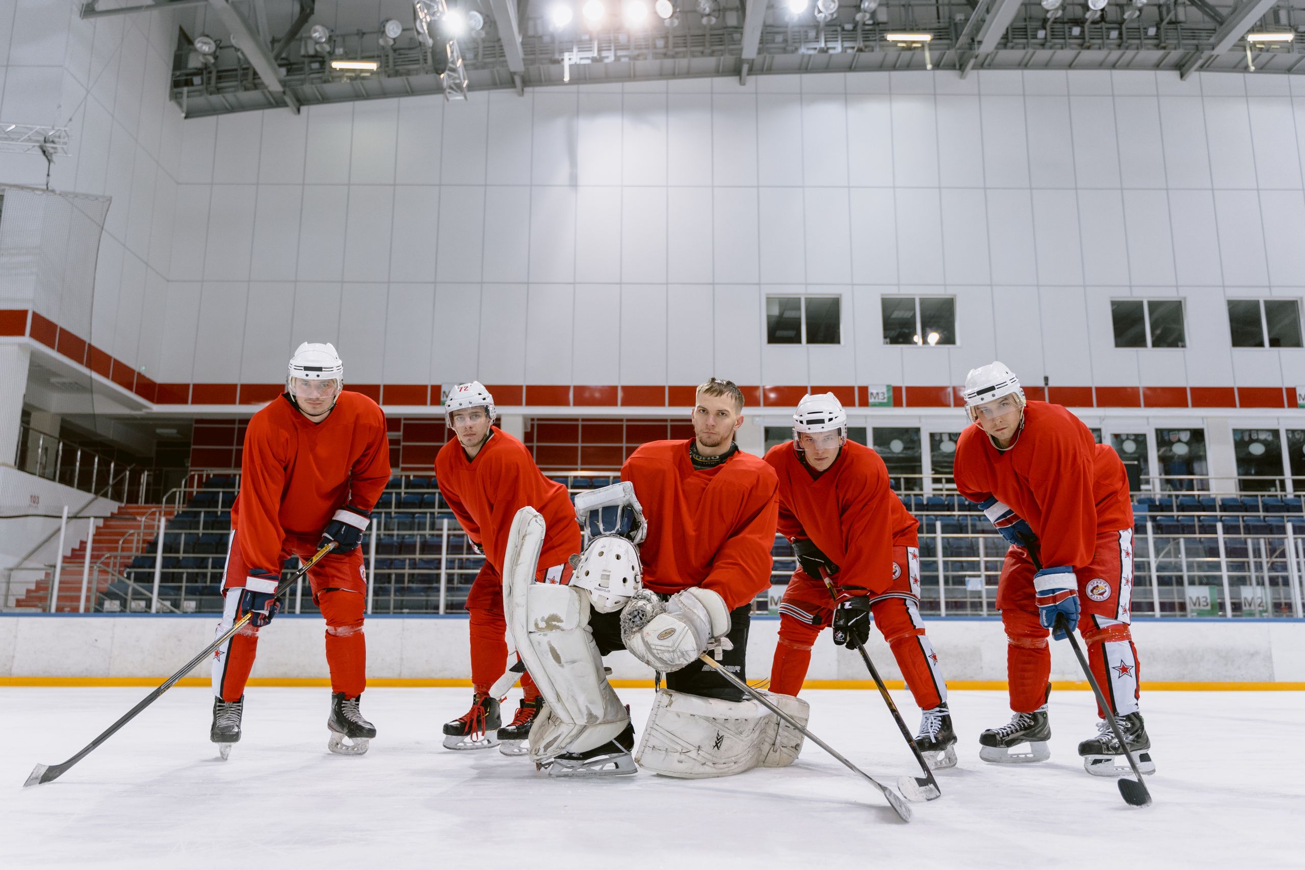 Five male ice hockey players in red uniforms, posing on an indoor rink with hockey sticks and gear.