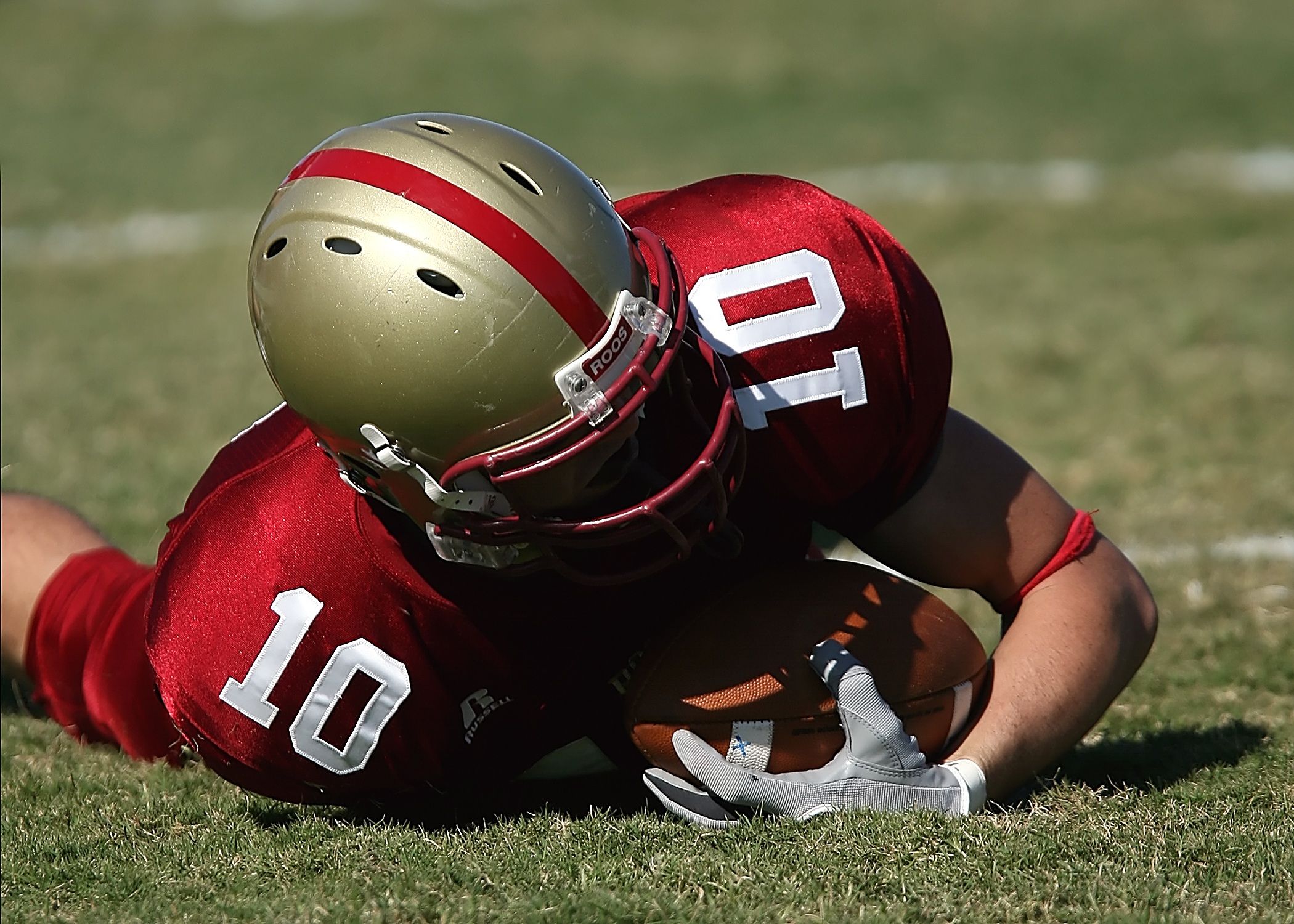 American football player wearing red uniform holding ball on grassy field during day game.