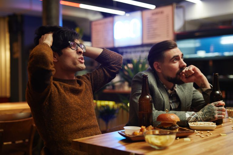 Two men in a modern bar sharing drinks and snacks, expressing mixed feelings.