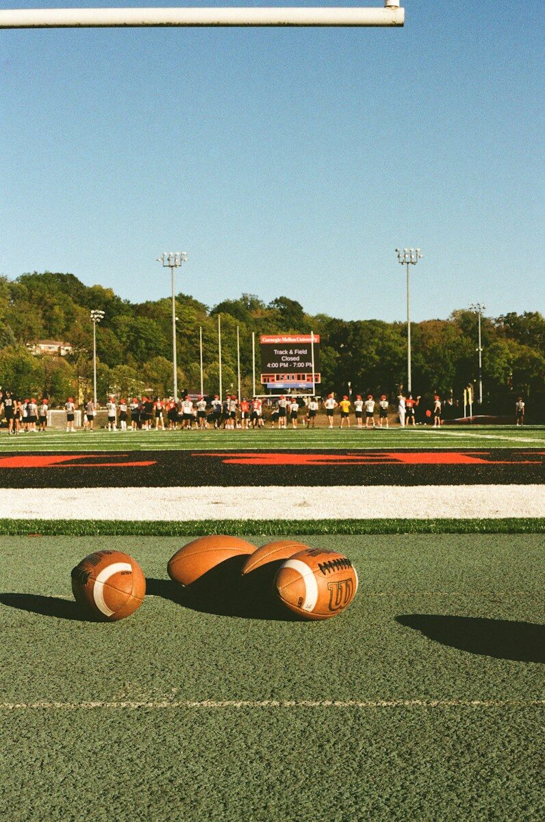 Three footballs on a green field with players in background