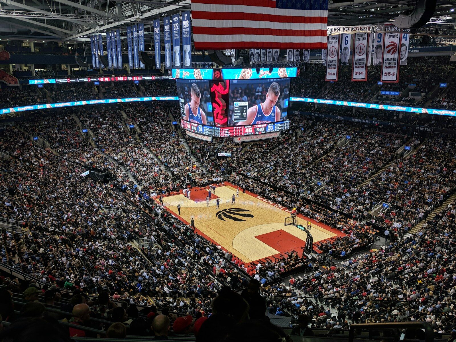 A crowded basketball arena with a large American flag hanging above the court